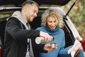 femme-blonde-bouclee-homme-assis-dans-coffre-voiture-dans-foret-automne-tasse-thermo